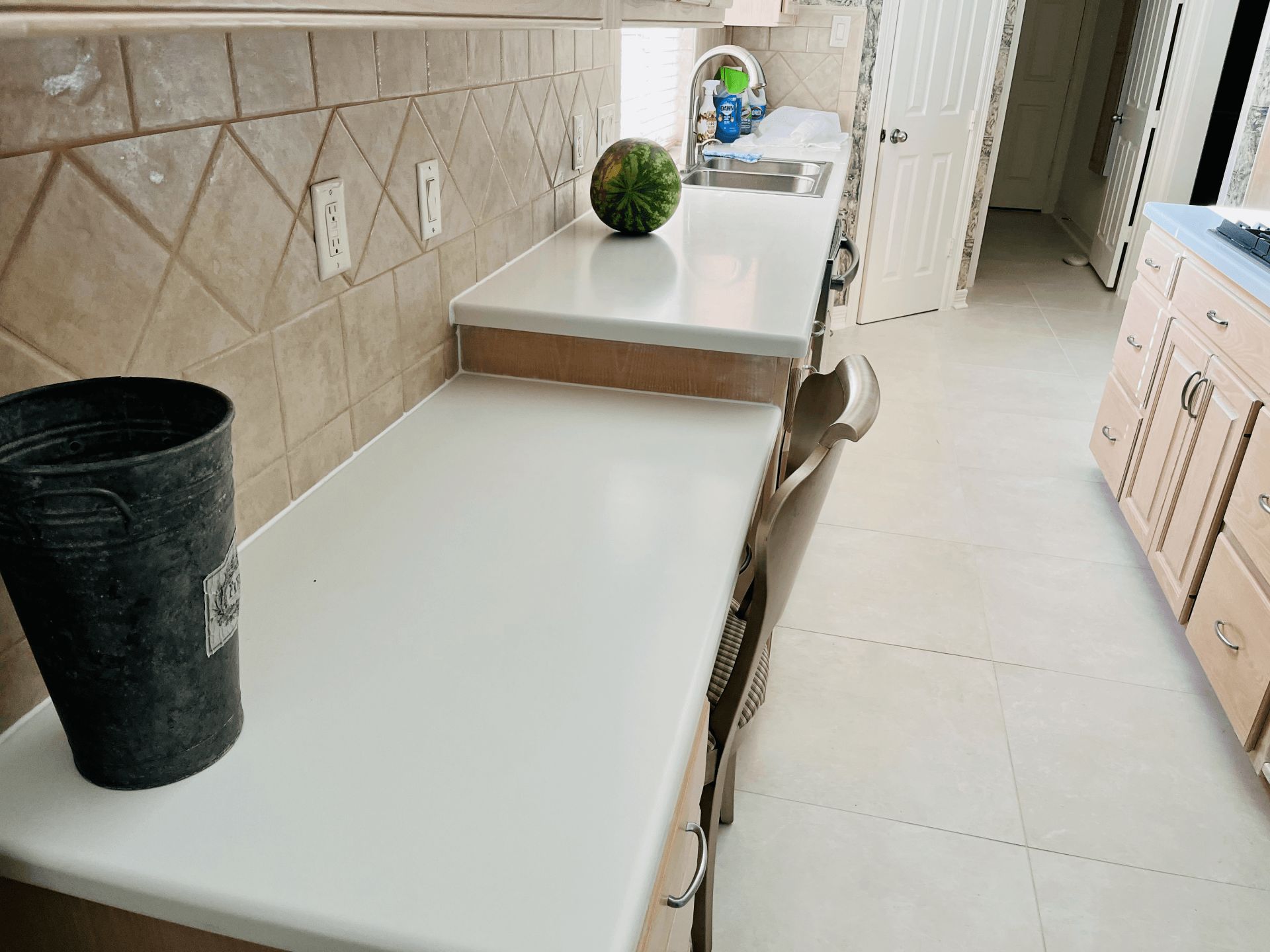 A kitchen countertop with a watermelon, a metal bucket, and a sink, next to light-colored cabinets and tiled flooring.