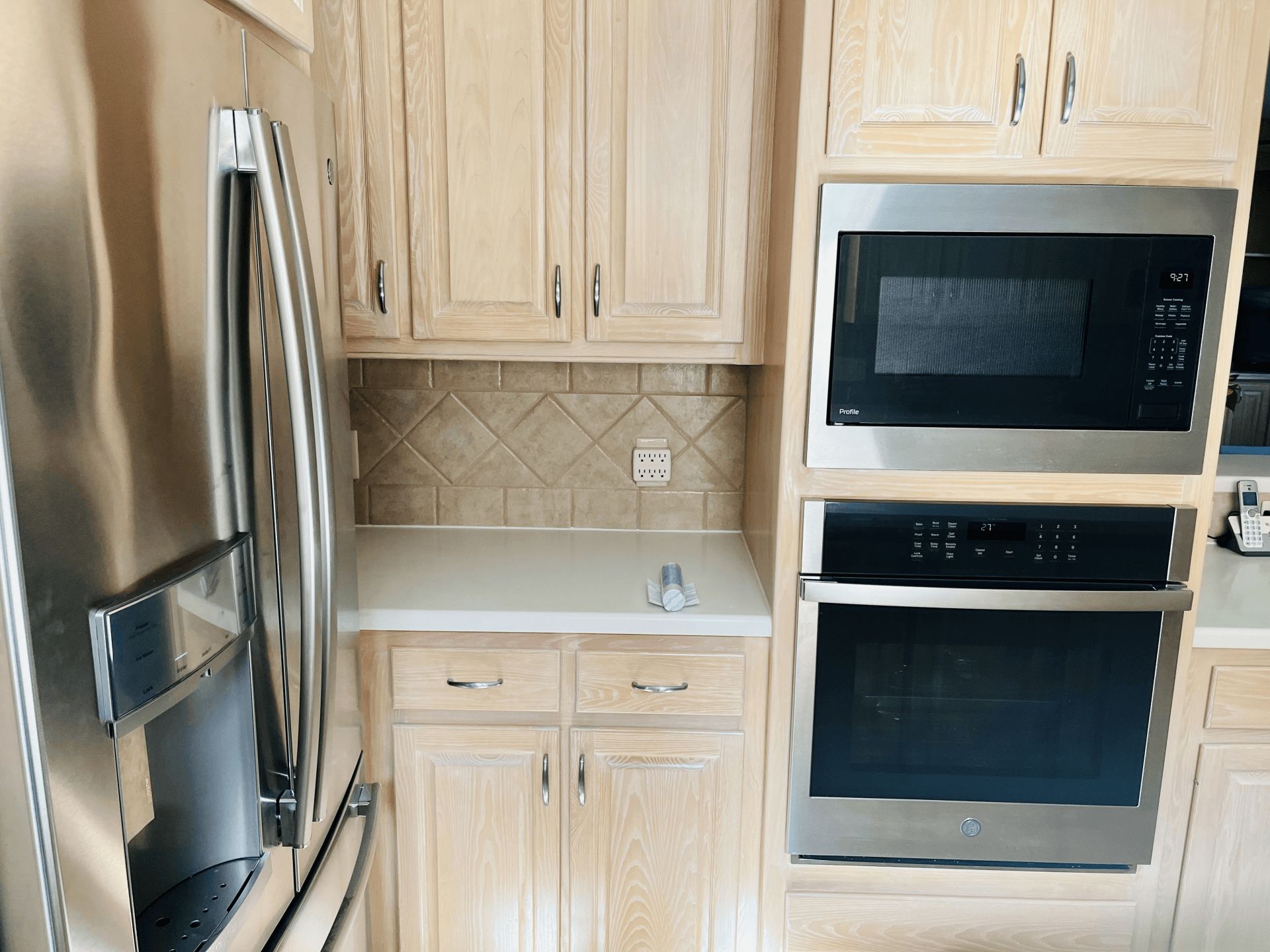 Stainless steel refrigerator, microwave, and wall oven in a kitchen with light wood cabinets and a tiled backsplash.