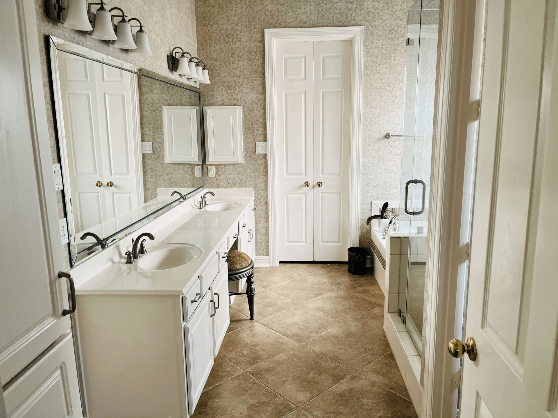 A bright bathroom with white vanity cabinets, a large mirror, beige textured wallpaper, and a shower-tub combo.