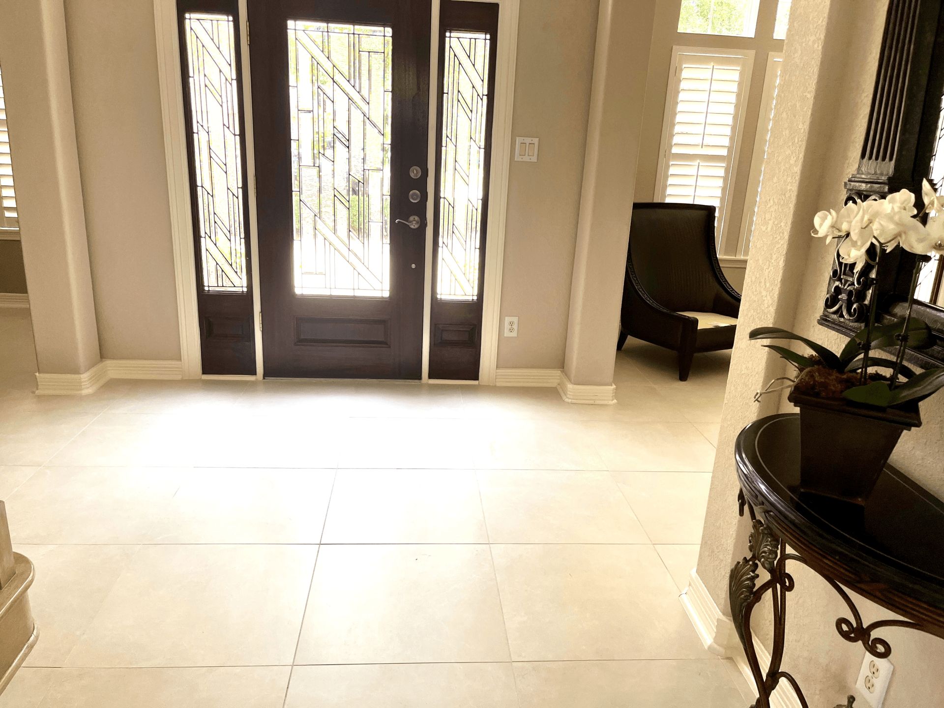 A bright entryway with beige tiled floors, a dark wooden front door with glass panels, and an ornate side console table.