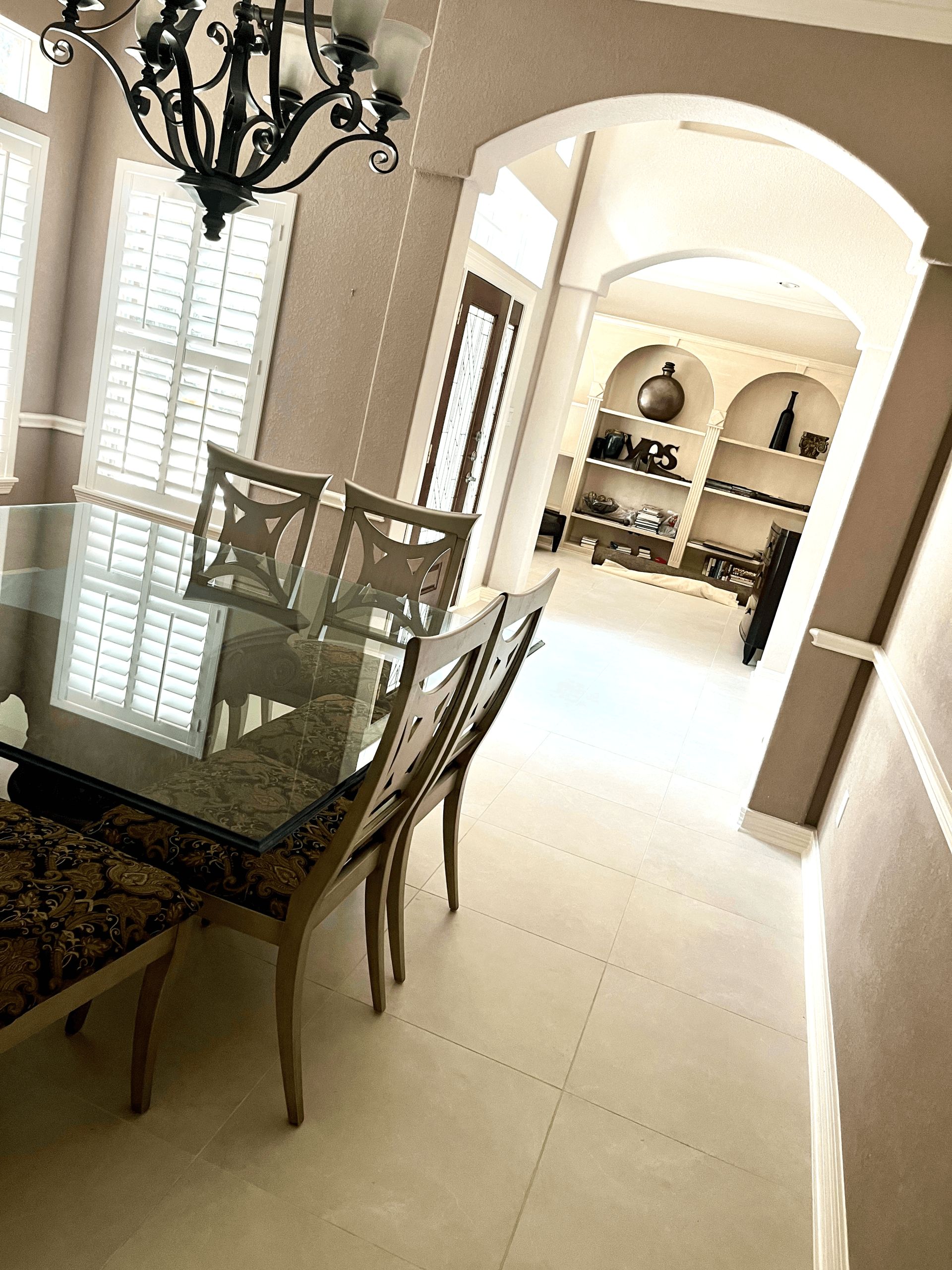 A dining area with a glass table, patterned chairs, and a chandelier, leading through an archway to a display shelf.
