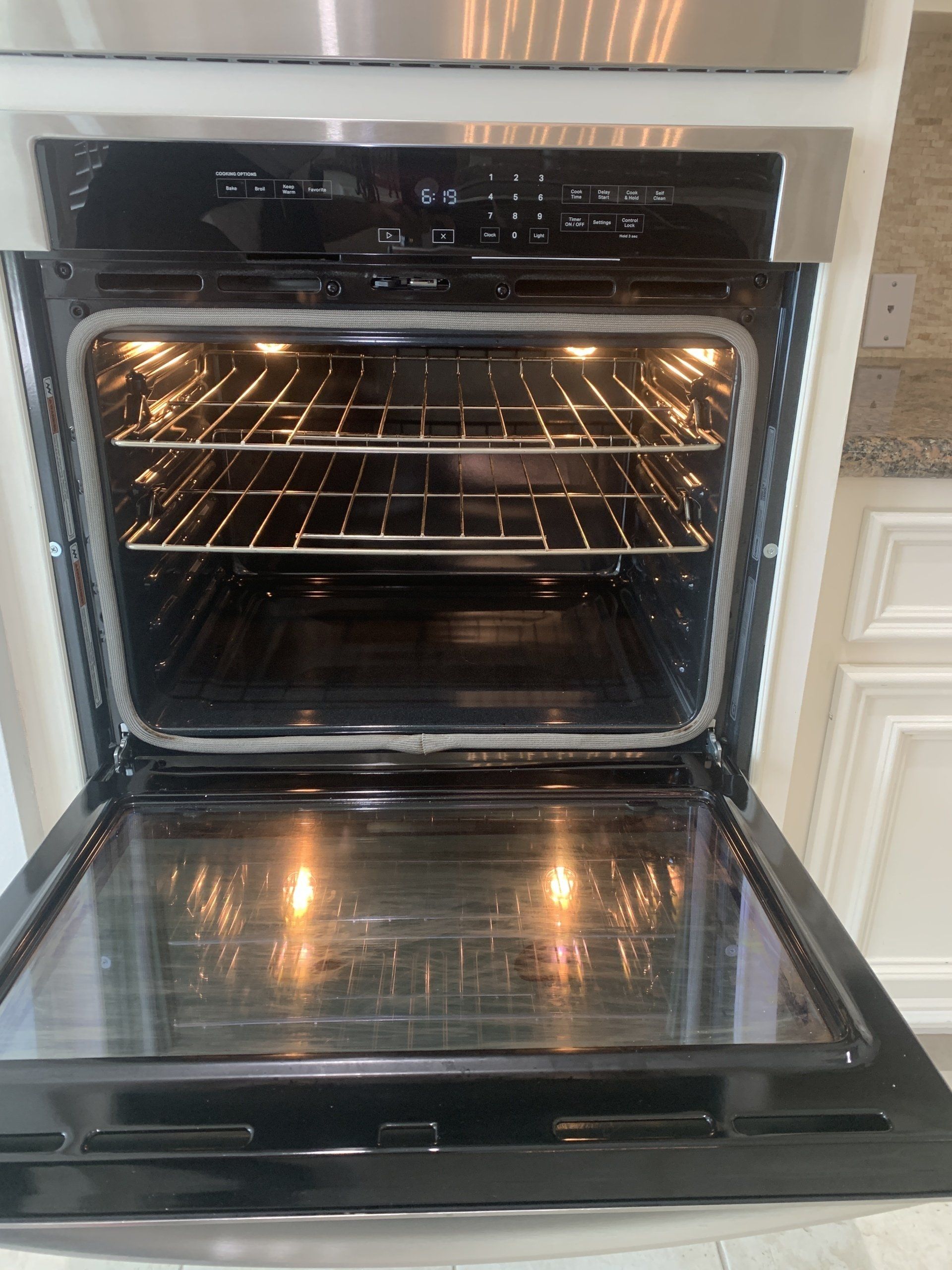 A built-in stainless steel oven with the door open, revealing a clean interior with two racks and bright lights.