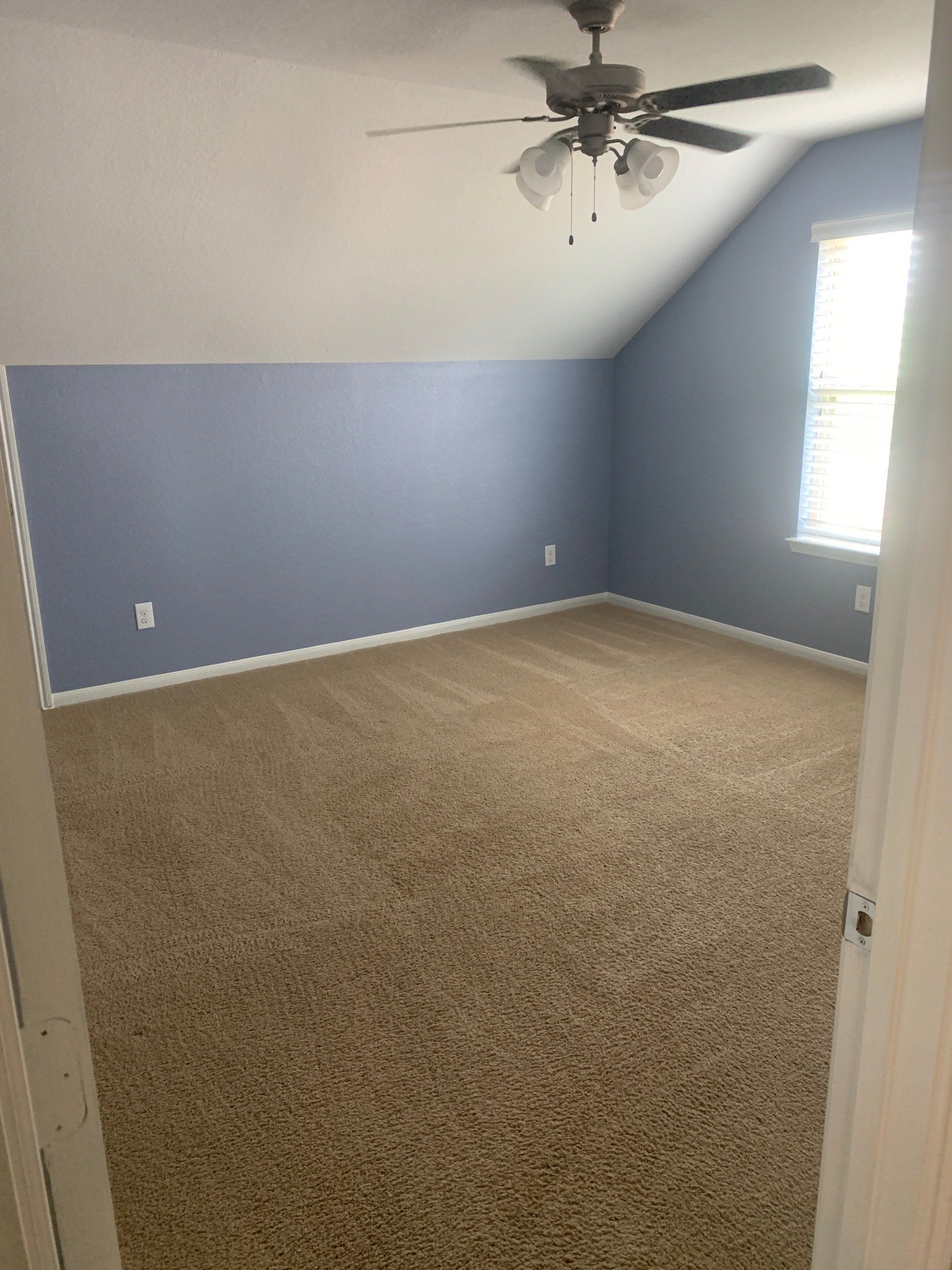 Empty room with sloped light blue walls, tan carpet, and a ceiling fan near a window with white blinds.