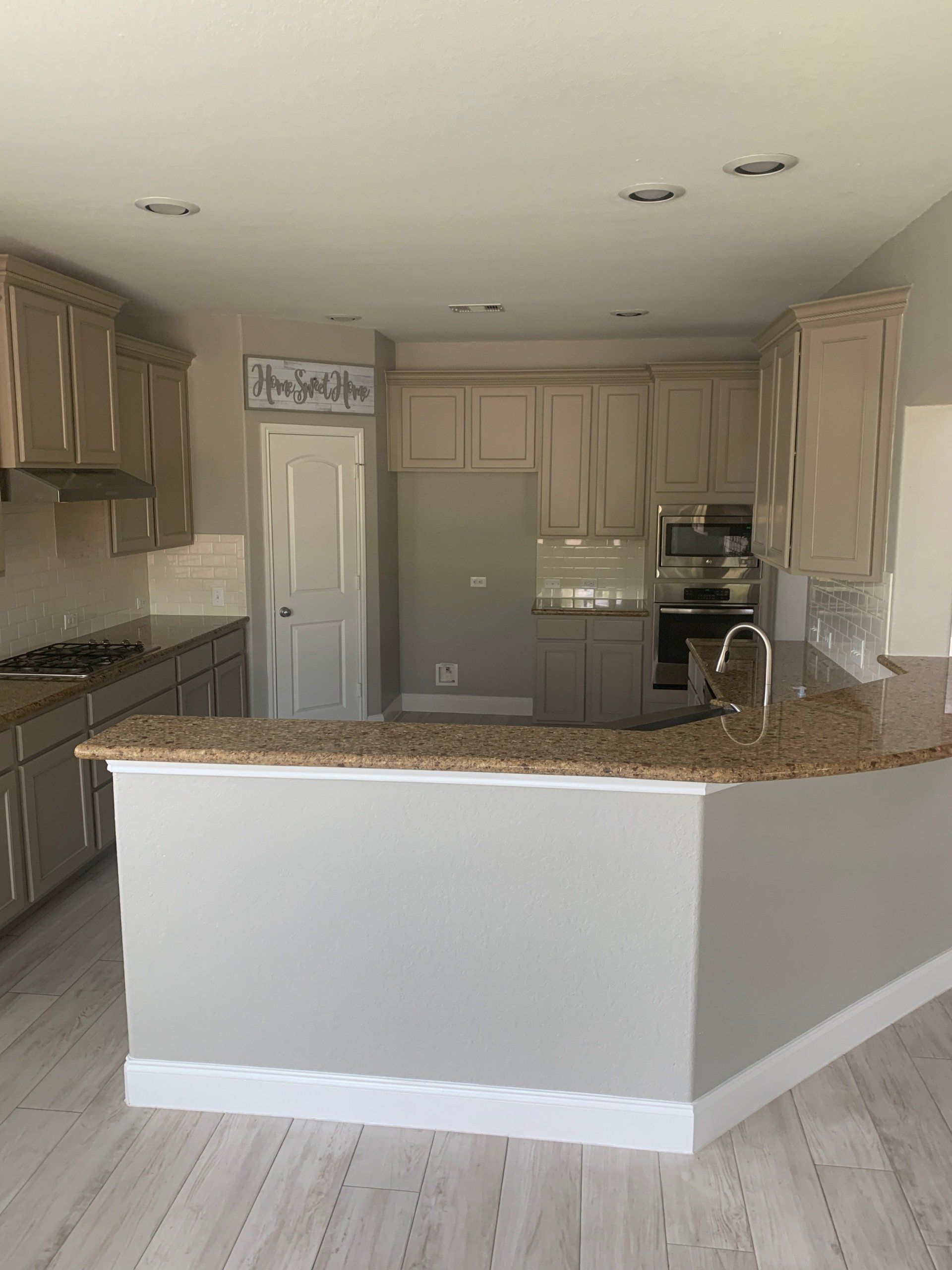 A modern kitchen featuring light taupe cabinets, granite countertops, an L-shaped island, and light-toned wood flooring.