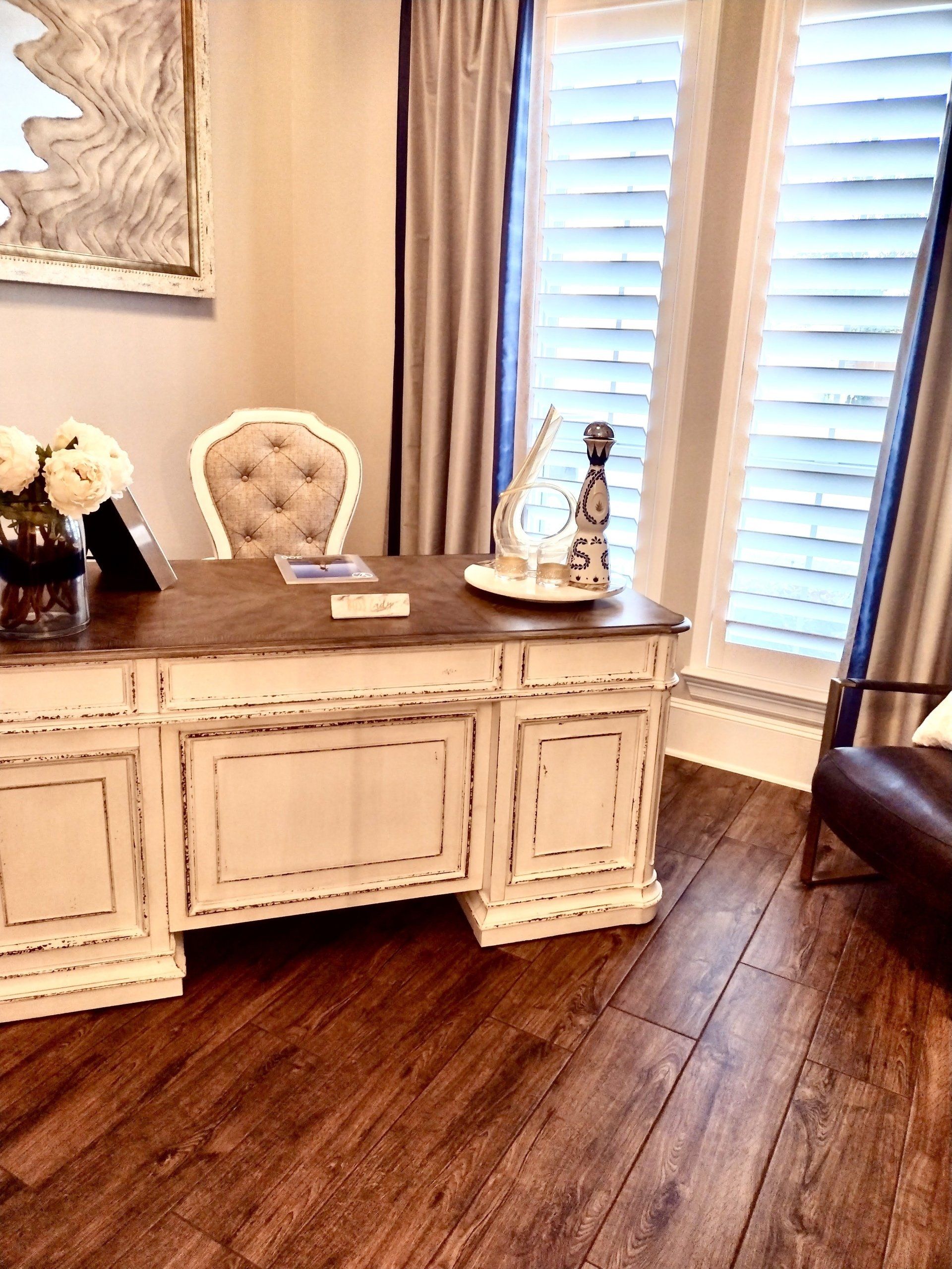 A distressed white desk with a tufted chair sits on wood floors in a sunlit room with window shutters and curtains.