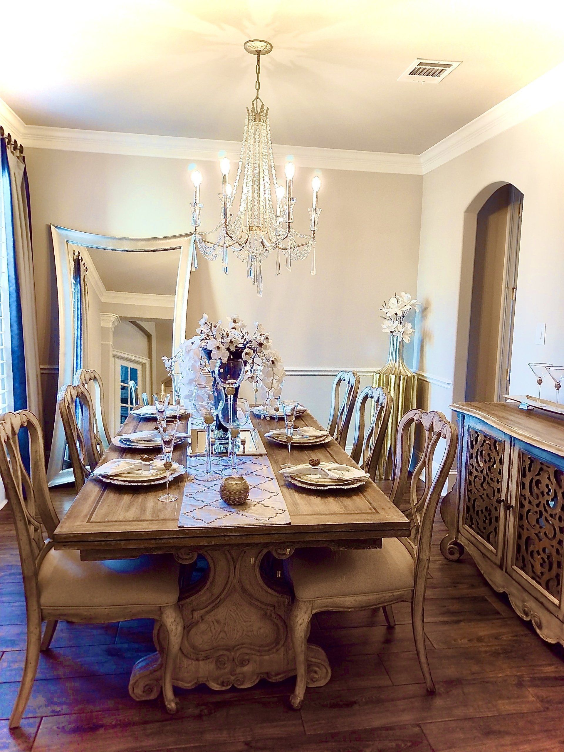 A dining room with a rustic wooden table set for a meal, a crystal chandelier, and a large ornate mirror.