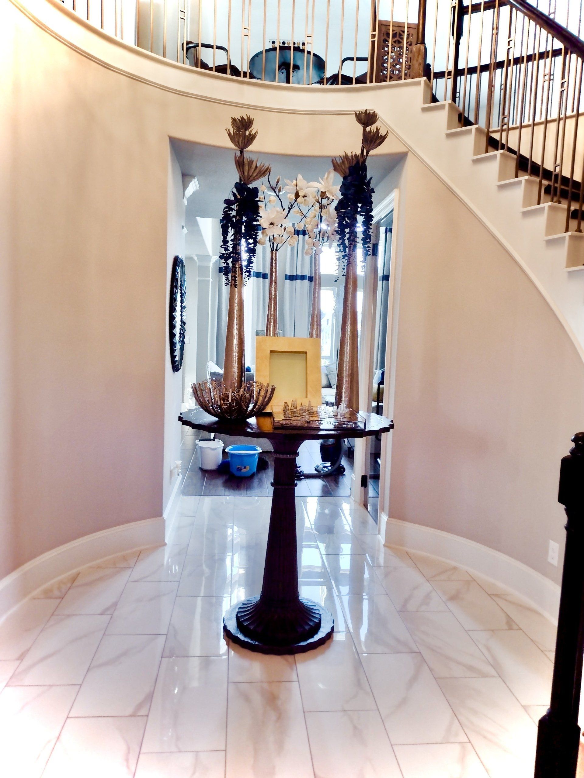 A curved hallway with light walls, a marble floor, a central pedestal table, and a grand staircase in the background.