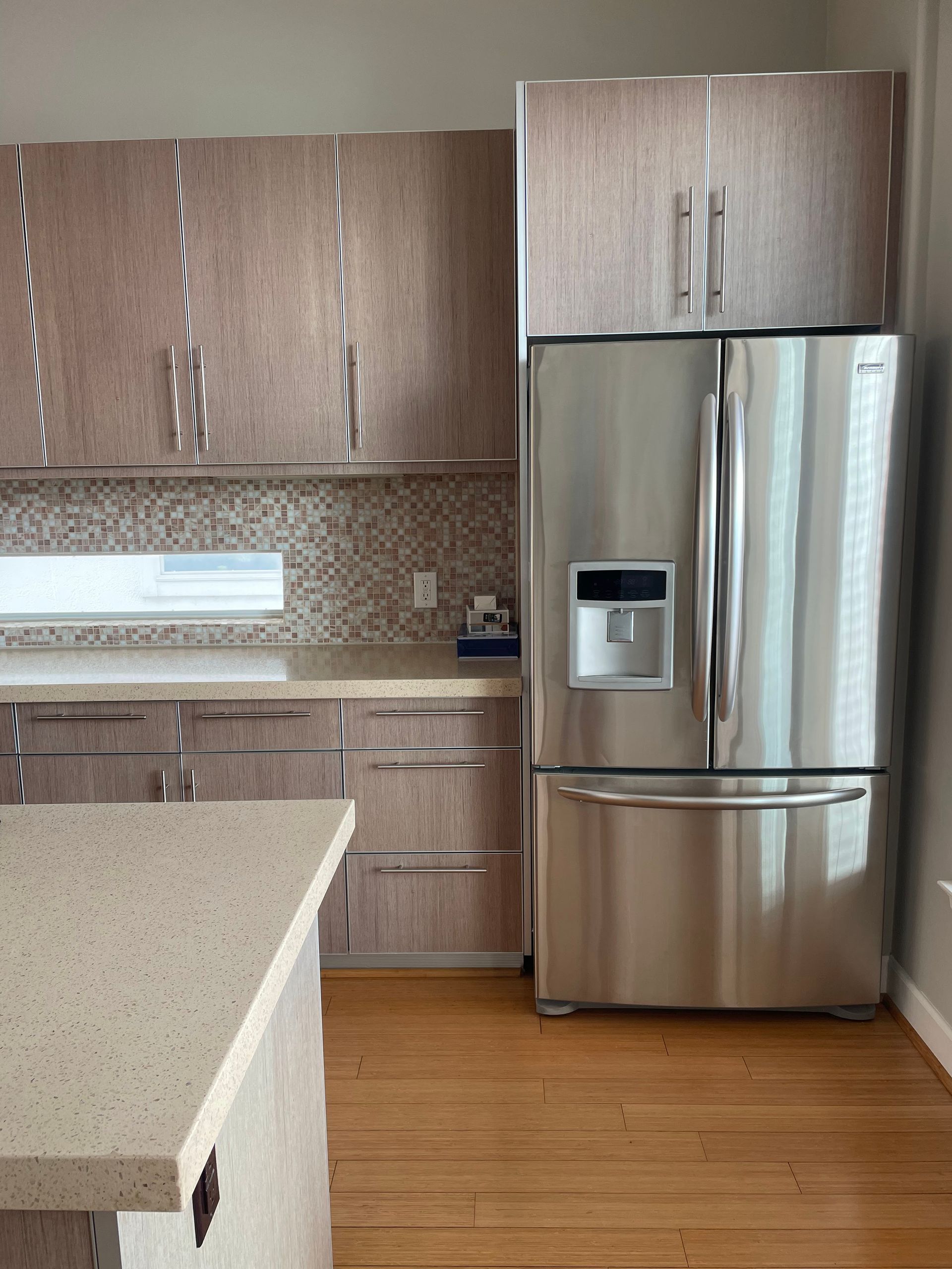 A modern kitchen featuring wood-grain cabinets, a stainless steel refrigerator, speckled countertops, and wood floors.