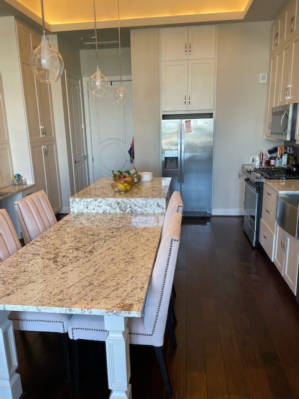 A kitchen with granite counters, light-colored upholstered chairs, pendant lights, and stainless steel appliances.