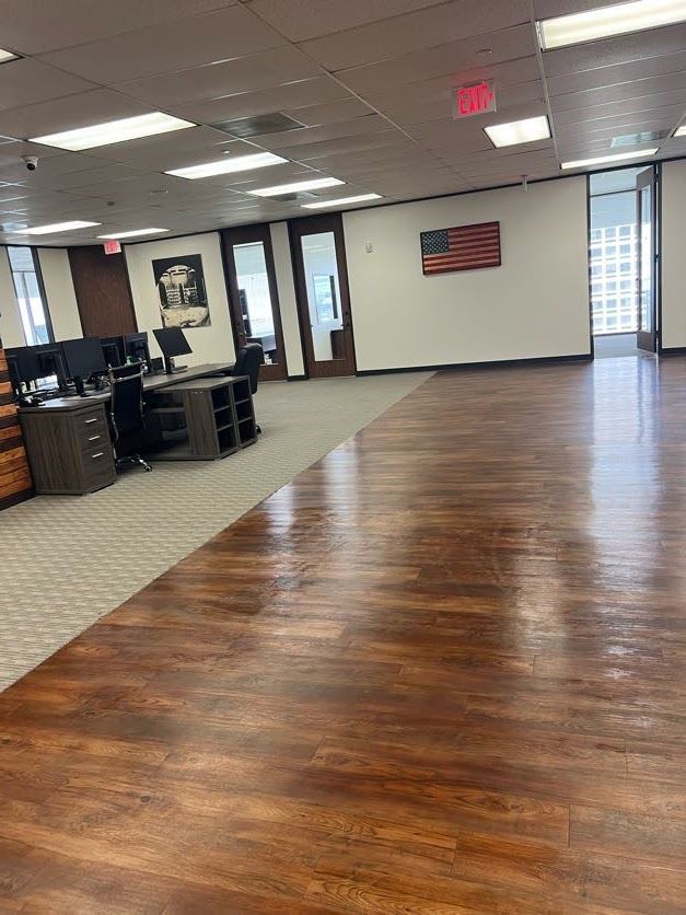 An open-plan office features wood-look floors, beige carpeting, a row of desks, glass-paneled doors, and a US flag on wall.