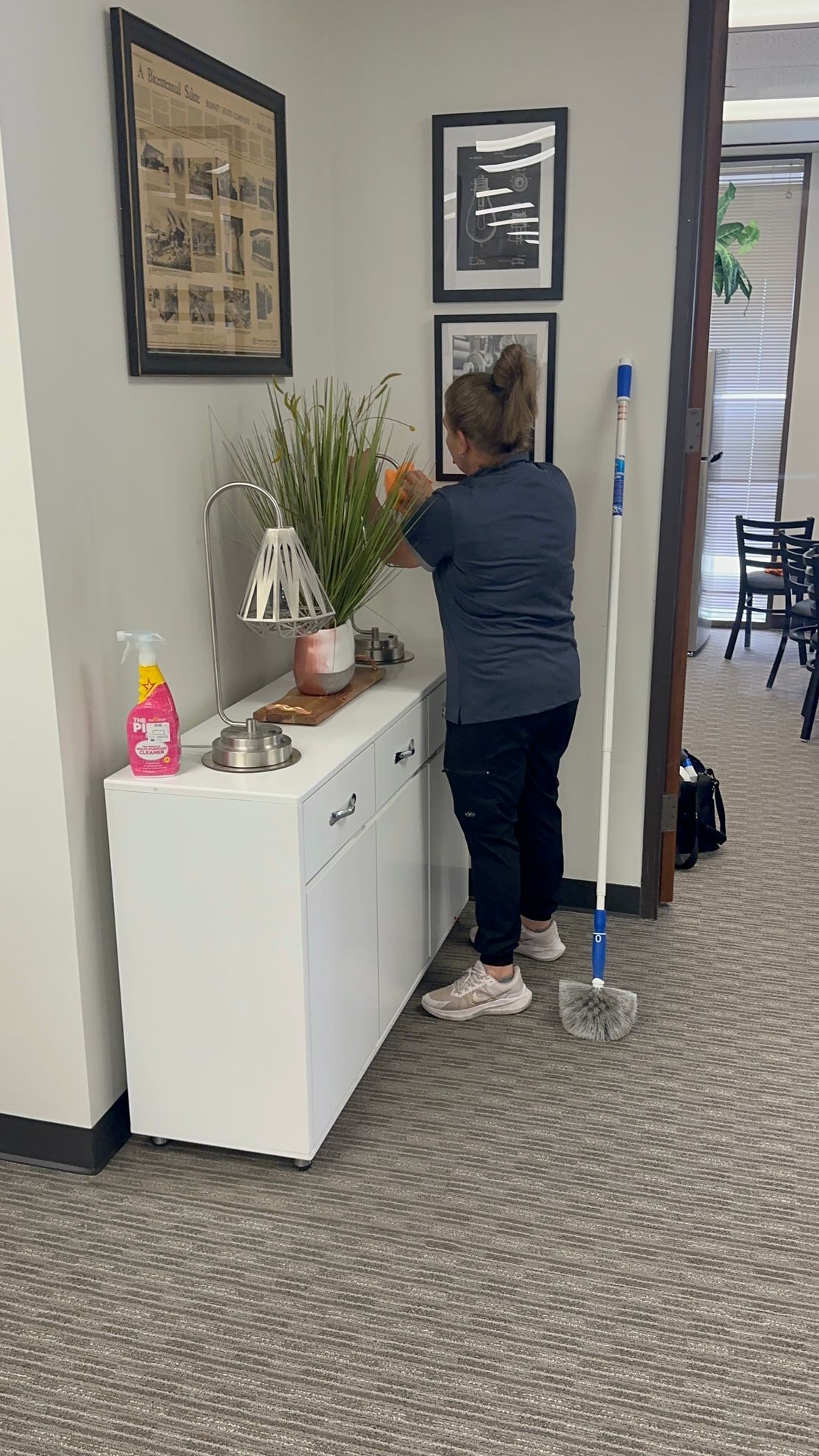 A person in a blue shirt cleans a wall above a white cabinet in an office, with a mop standing nearby.