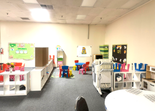 A brightly lit classroom with low white shelving, small tables and chairs, and colorful wall decor for children.