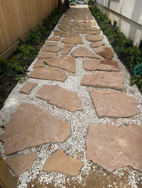 Path of irregularly shaped brown stones through gravel, between a wooden fence and a building.