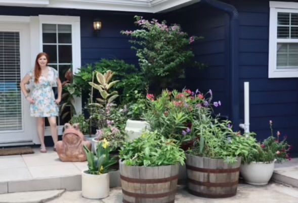 Woman stands by a lush garden of potted plants, in front of a blue house with white trim and doors.