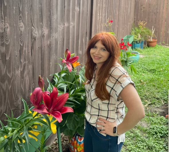 Woman with red hair and plaid shirt near blooming red lilies in a garden.
