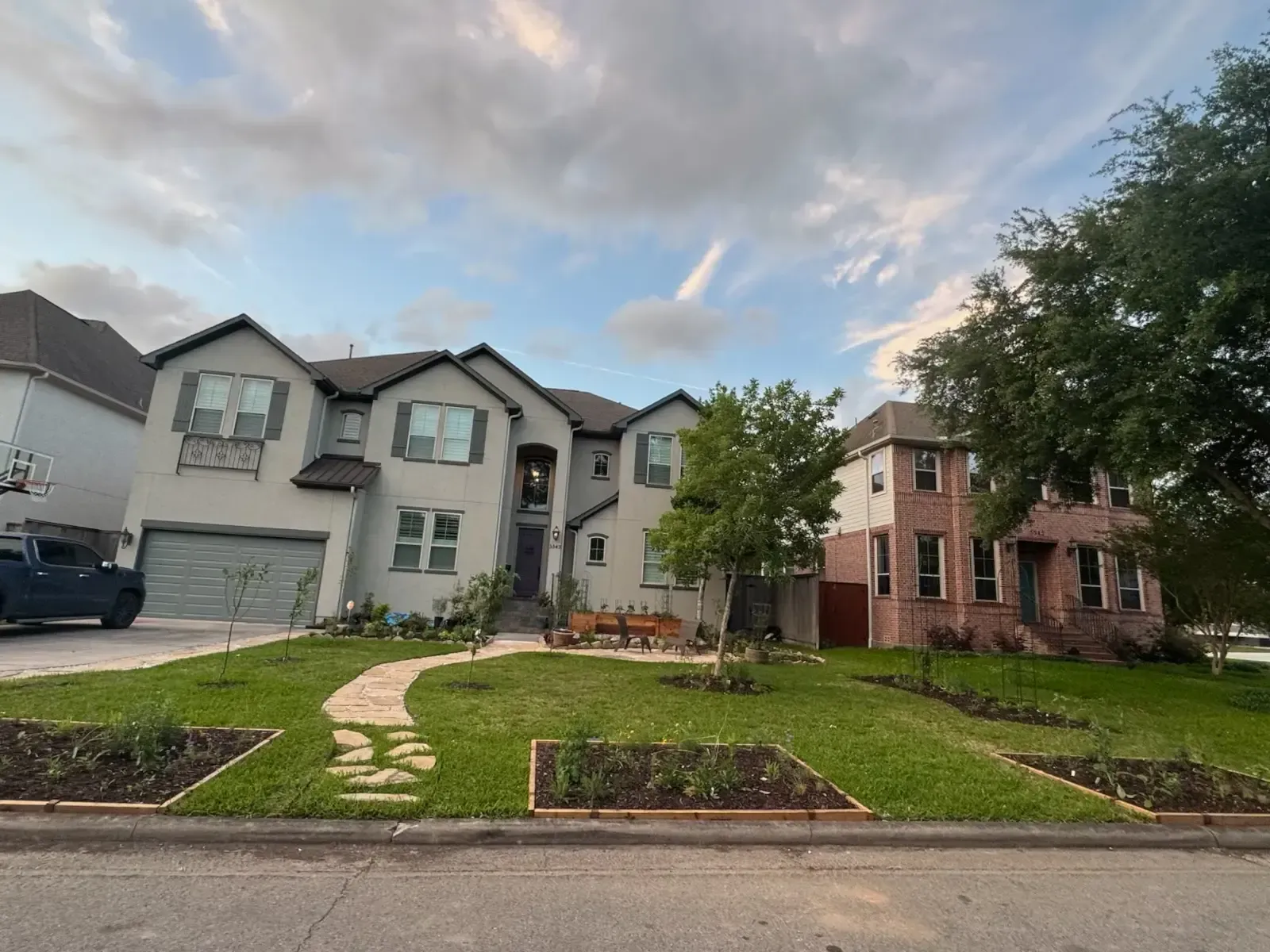 Two-story stucco house with stone pathway and garden beds. Cloudy sky, car parked in driveway, neighbor's brick house visible.
