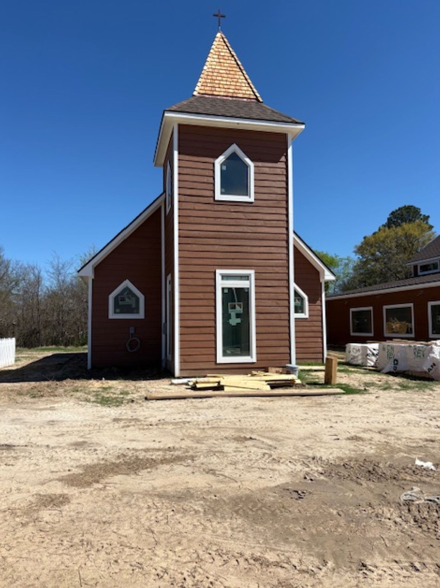 A newly constructed brown church with a tall steeple and white-trimmed windows under a clear blue sky.