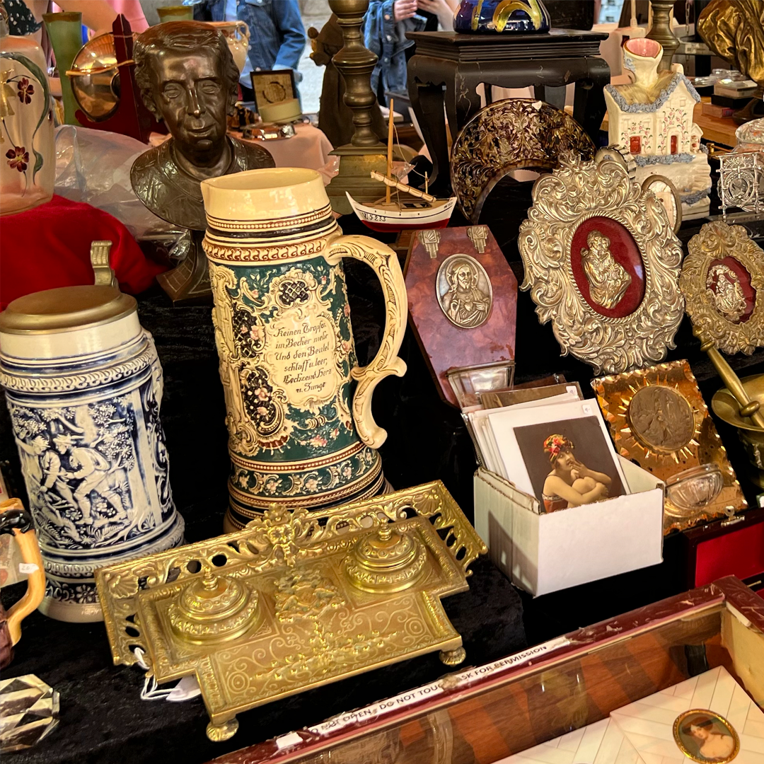 An assortment of antique items, including beer steins, a brass inkwell, and decorative framed portraits, on a table.