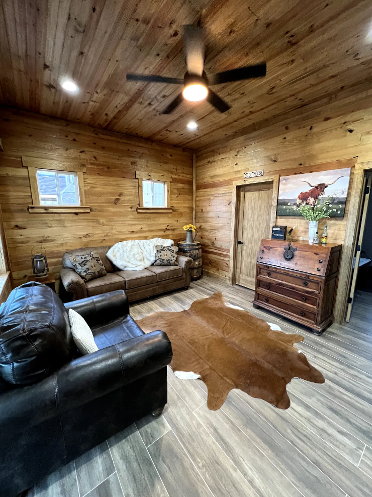 A rustic living room with wood paneled walls, two leather sofas, a cowhide rug, and a wooden secretary desk.