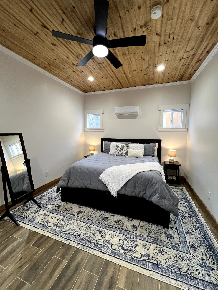 A modern bedroom featuring a bed with gray bedding, a wood-paneled ceiling, a ceiling fan, and a patterned area rug.