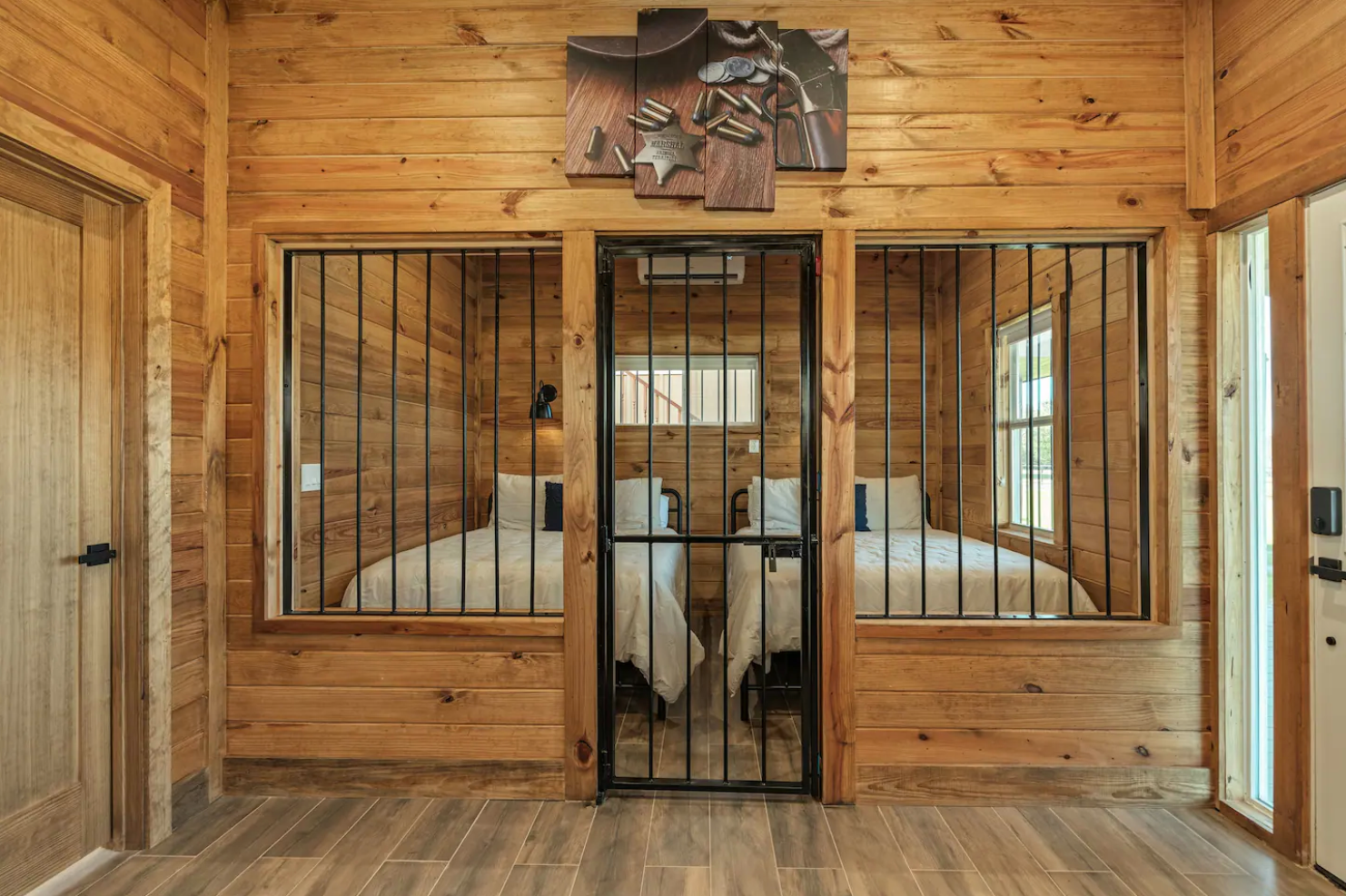 A room with wooden walls and flooring features a central barred doorway flanked by windows looking into a bunk room.