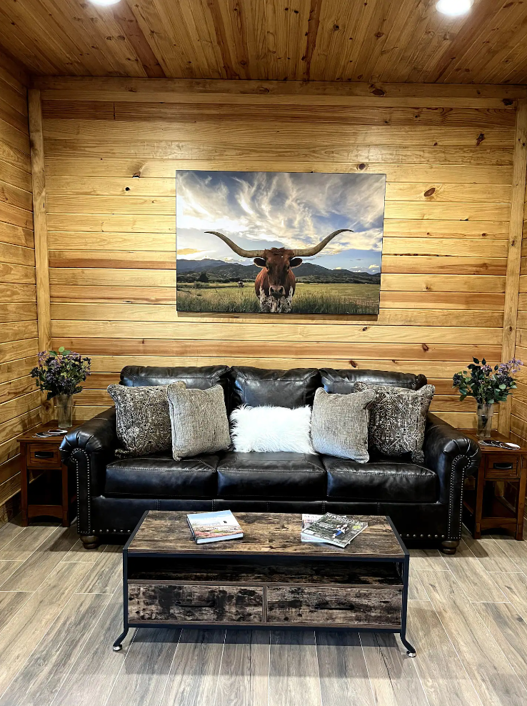 A rustic room with wood walls, a leather sofa, a coffee table, and a wall print of a longhorn steer in a field.