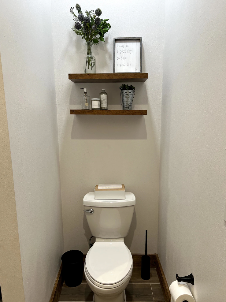 A white toilet in a small bathroom with two wooden floating shelves holding decor, a plant, and a framed sign.