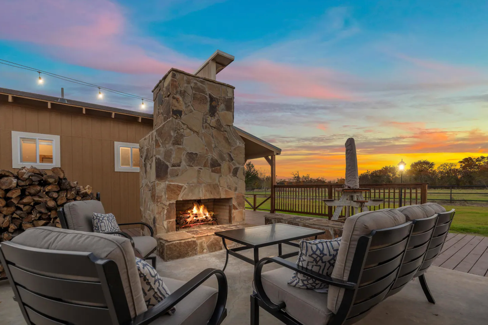 A stone outdoor fireplace on a patio with cozy chairs at sunset, featuring a pile of firewood and a wooden deck.