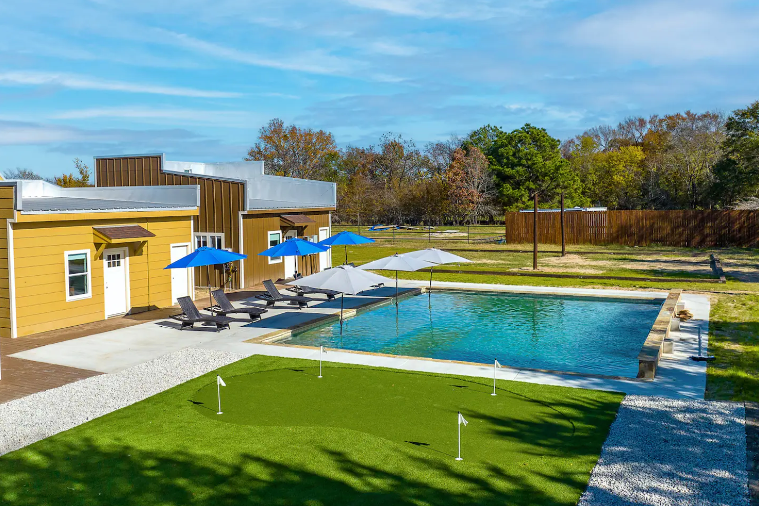 A yellow structure with an adjacent swimming pool, umbrellas, and a small putting green under a bright blue sky.