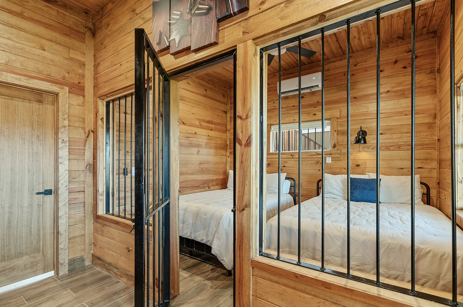 A bedroom with two beds seen through a black iron-barred gate and window wall in a room with light wood paneling.