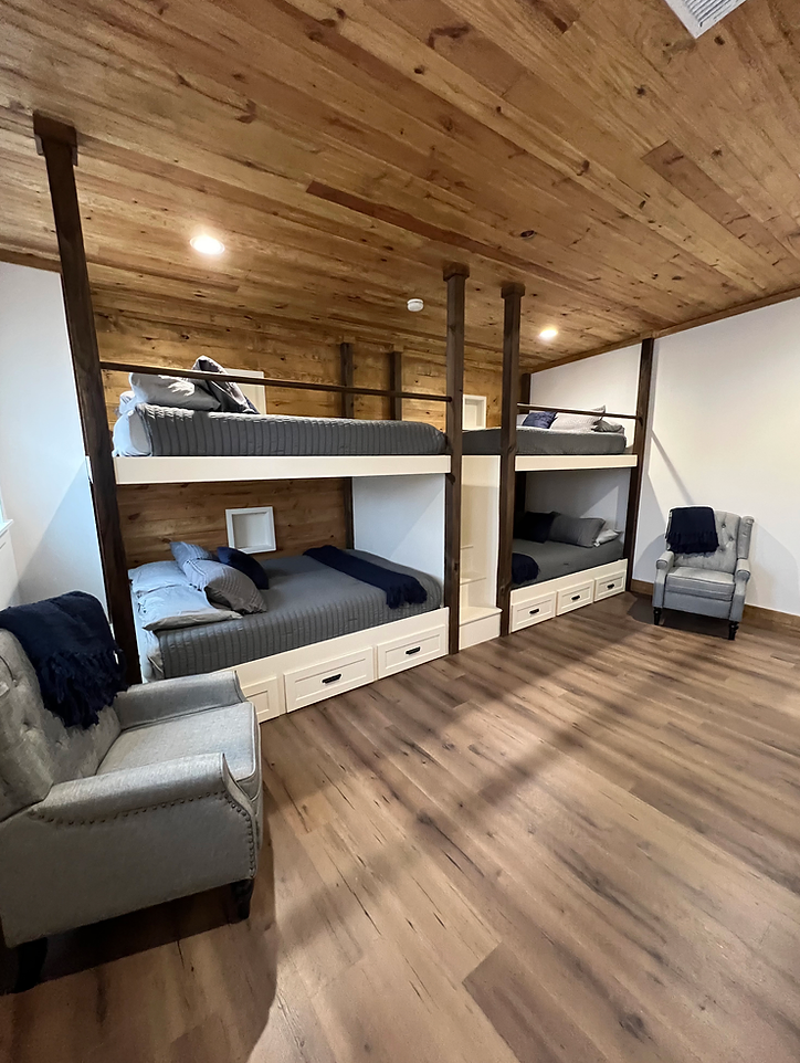 A room with two sets of wood-framed bunk beds, grey bedding, and two grey armchairs on light wood floors.