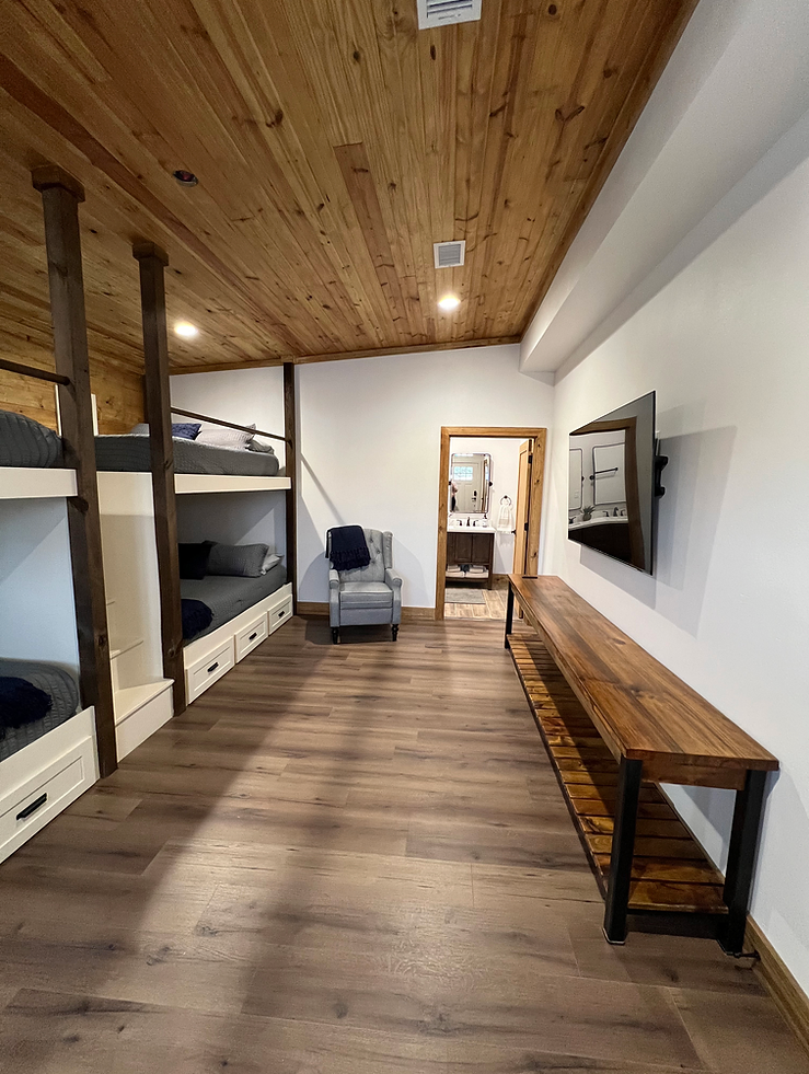 A bunk room with wood-paneled ceiling, white walls, built-in wooden bunk beds, a grey armchair, and a wall-mounted TV.