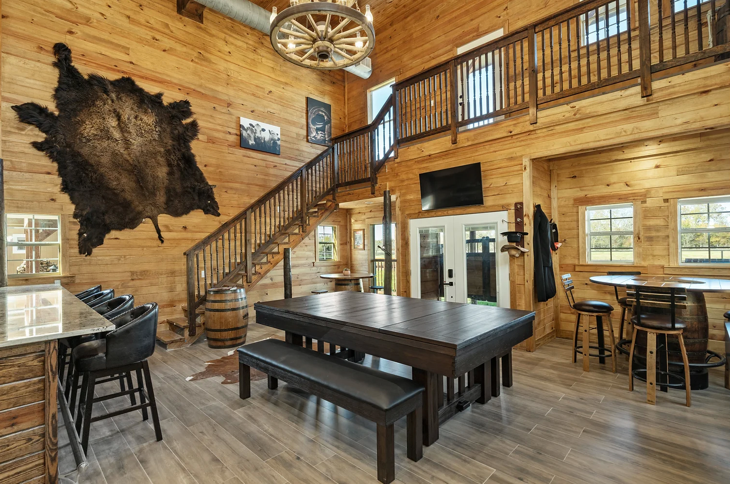 A rustic game room with wood walls, a pool table, stools, and a staircase leading to an upstairs balcony.