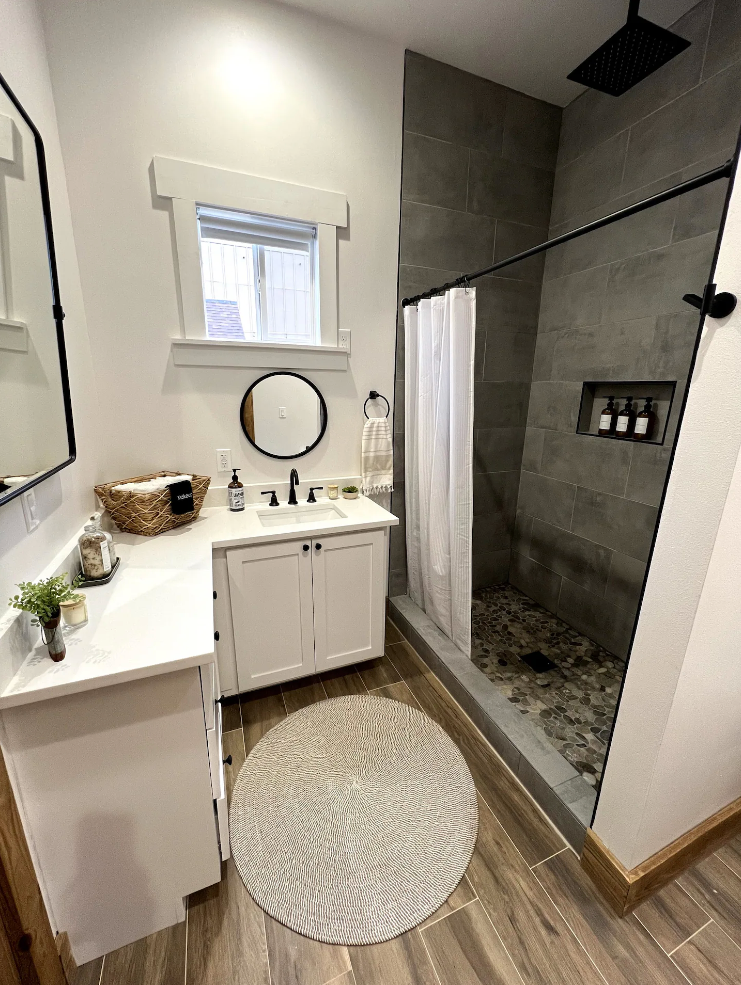 A modern bathroom features a white vanity, a round wall mirror, a woven basket, and a walk-in shower with gray tile.