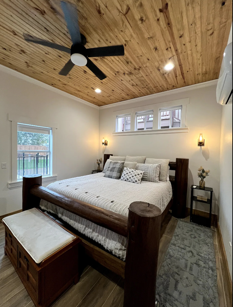 A cozy bedroom featuring a rustic log bed, wooden plank ceiling, ceiling fan, white bedding, and a wooden storage bench.