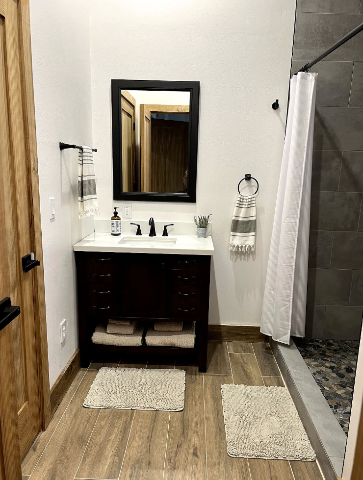 A bathroom featuring a dark wood vanity with a white countertop, a black-framed mirror, and a walk-in shower with stone tile.