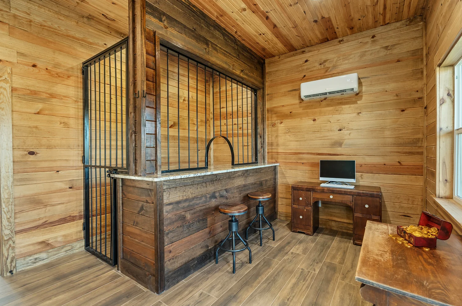 A rustic room with wood walls, a bar with iron bars, two stools, a desk with a computer, and a wood table.