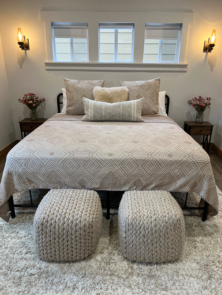 A bedroom features a bed with a patterned beige quilt, two ottomans at the foot, bedside tables, lamps, and a window.