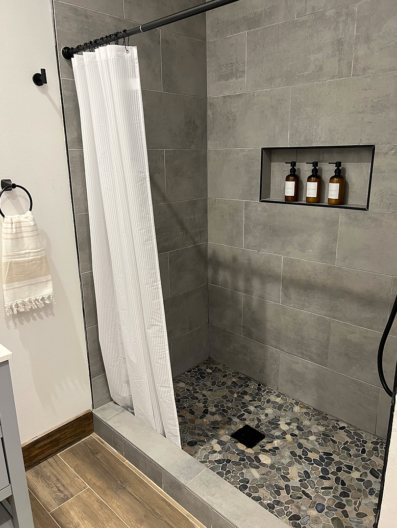 A walk-in shower with gray tiled walls, a pebble floor, a built-in recessed niche for bottles, and a white shower curtain.