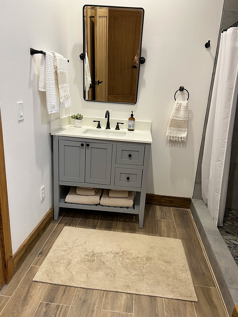 A bathroom vanity with a grey cabinet, white countertop, dark faucet, mirror, towels, and a rug on wood-look flooring.