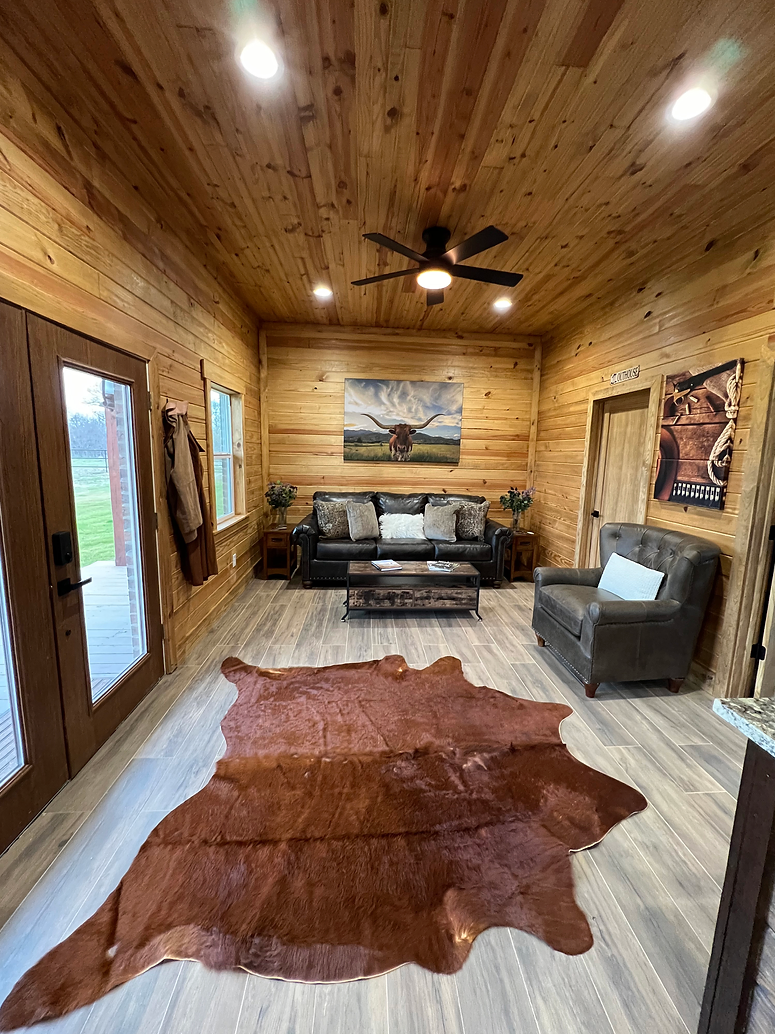 Rustic living room with wood paneled walls and ceiling, a dark leather sofa, armchair, and a large brown cowhide rug.