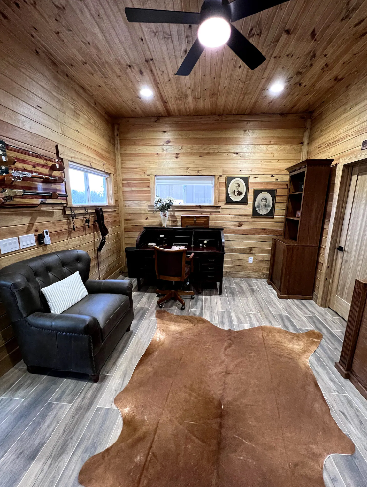 Rustic wood-paneled office with a leather sofa, desk, chair, cowhide rug, and wall-mounted decor under a ceiling fan.