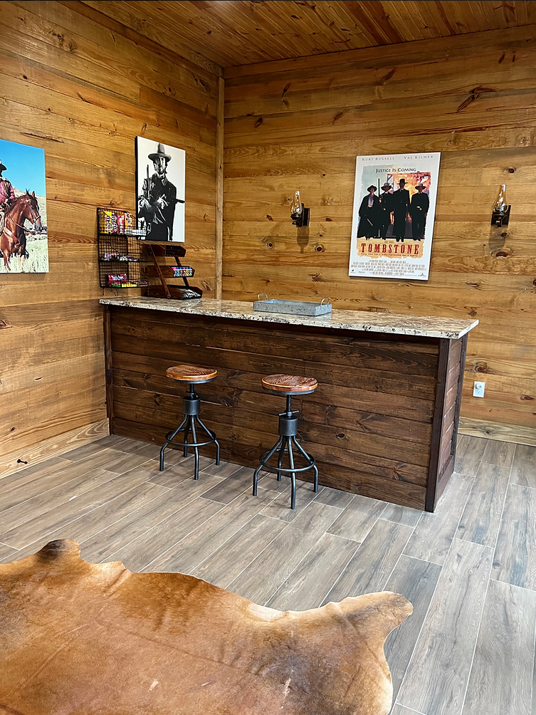 A rustic home bar with a granite countertop, two metal stools, wooden walls, Western-themed wall art, and a cowhide rug.
