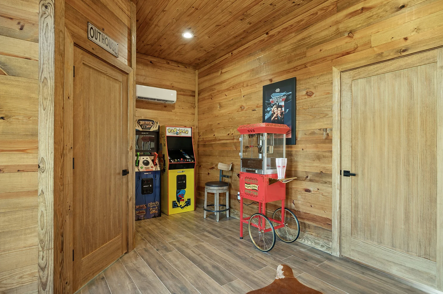 A wood-paneled game room features two arcade cabinets, a red popcorn machine, and a stool between two wooden doors.