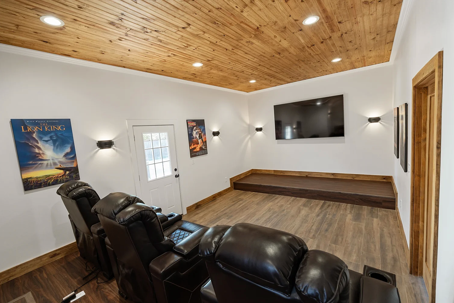A home theater room with black leather recliners, a large wall-mounted TV on a raised platform, and wooden ceiling planks.