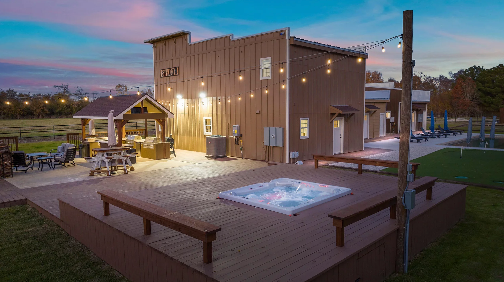 A wooden deck with a built-in hot tub, outdoor seating, and string lights next to a building at sunset.