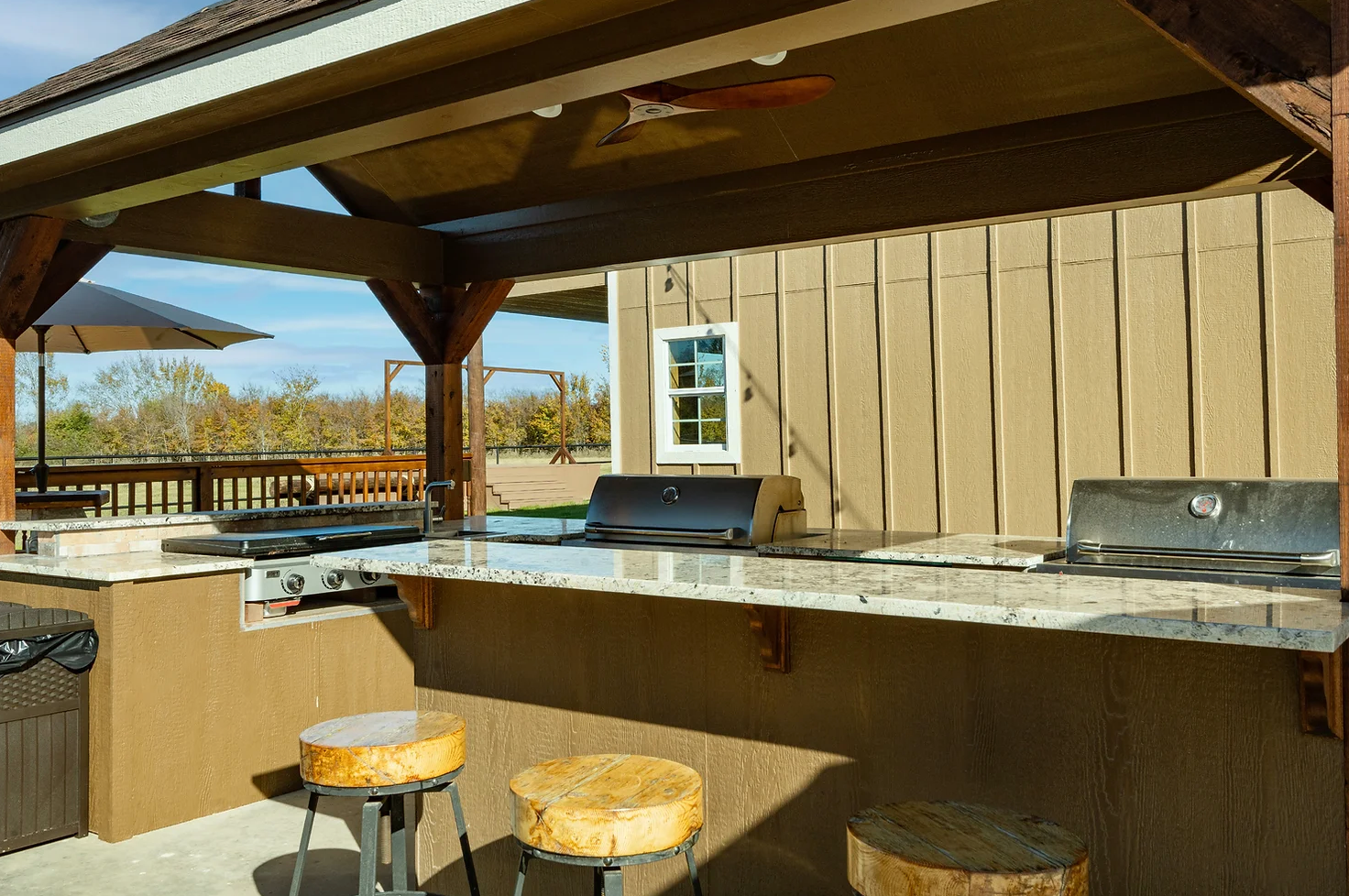 Outdoor kitchen under a wooden pavilion featuring a granite countertop, grill, and three bar stools on a concrete patio.