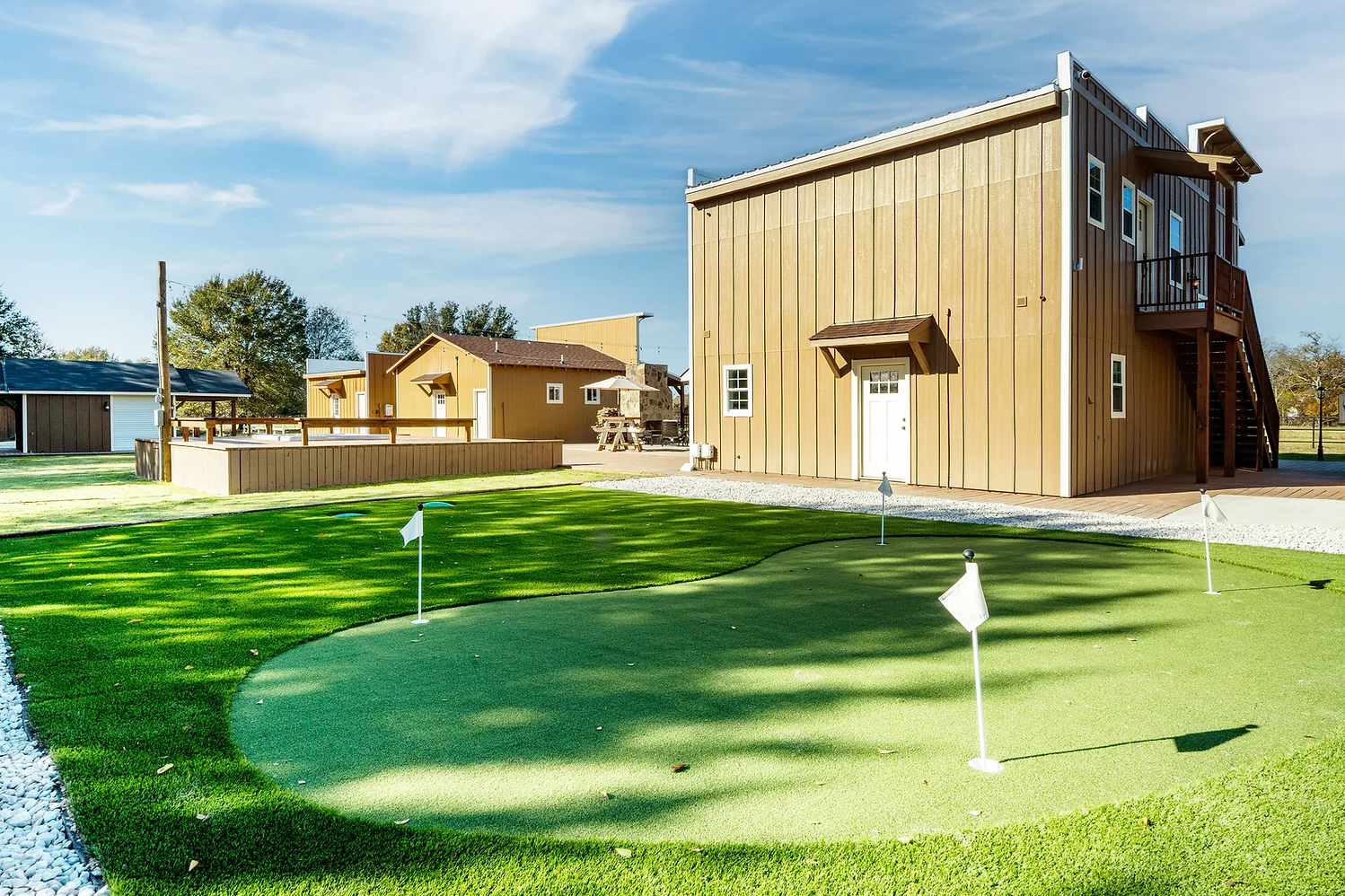 A putting green with golf flags sits in front of rustic wooden buildings under a blue sky.