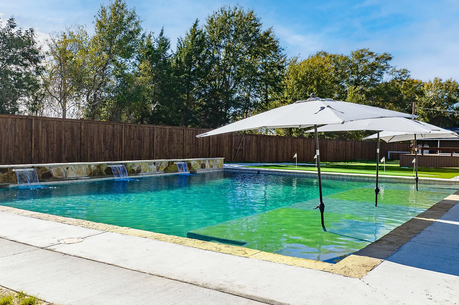 A backyard swimming pool with water features along a stone wall, blue water, and sun umbrellas under a bright sunny sky.