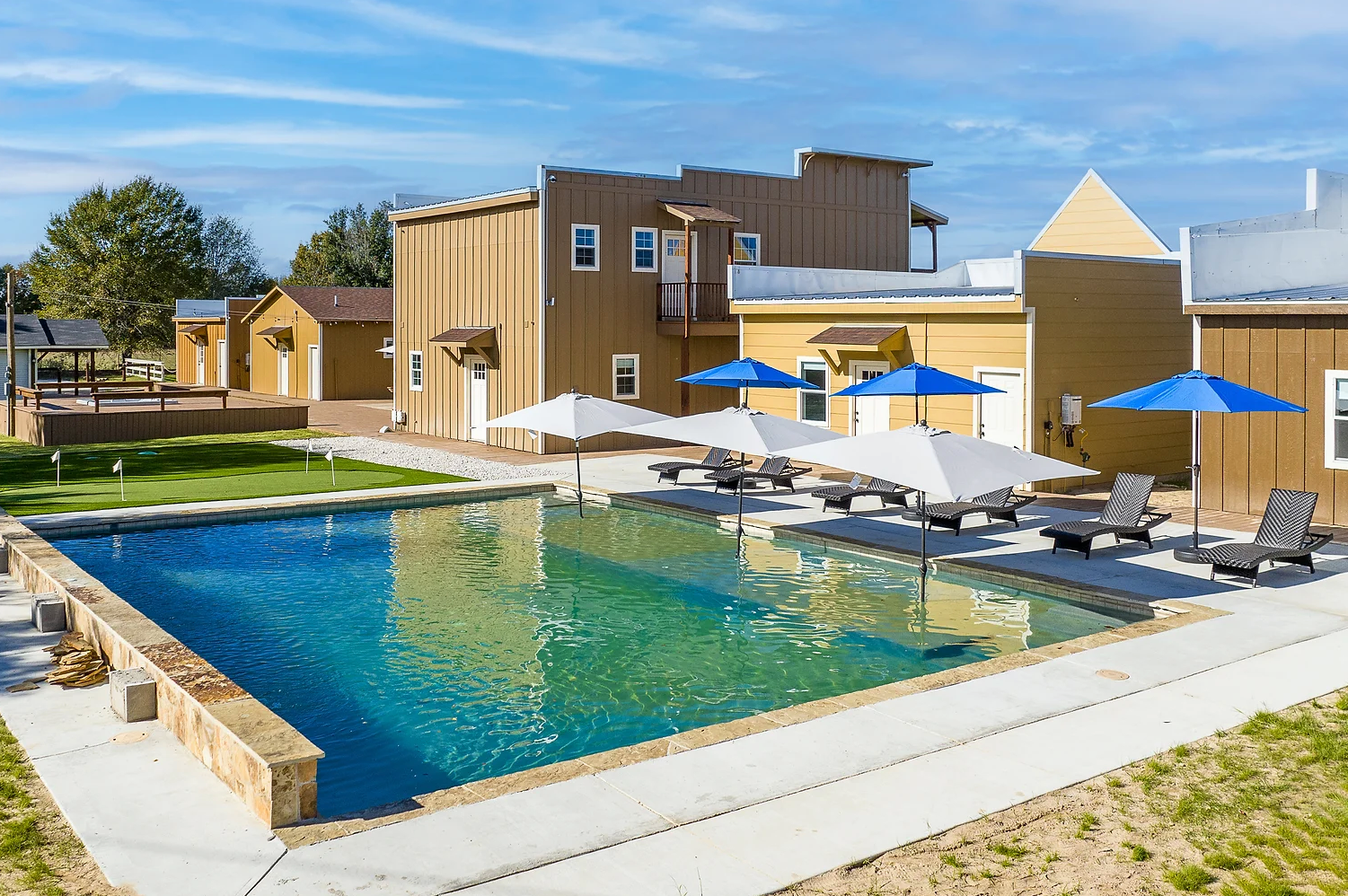 A rectangular swimming pool surrounded by a concrete deck with lounge chairs and blue umbrellas in front of rustic buildings.